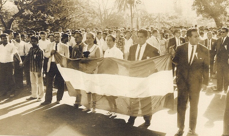 Seis personas portan una bandera larga, posando frente a una gran manifestación.
