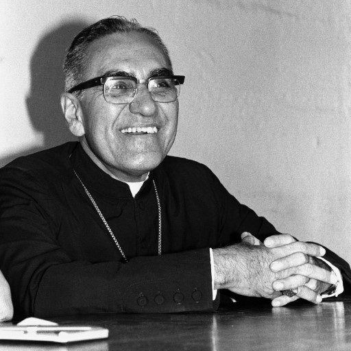 Una foto en blanco y negro del arzobispo Óscar Romero. Está sentado, con sotana y gafas, con las manos entrelazadas sobre la mesa. Sonríe, casi parece reírse ligeramente, mirando a alguien o algo fuera del encuadre de la fotografía.