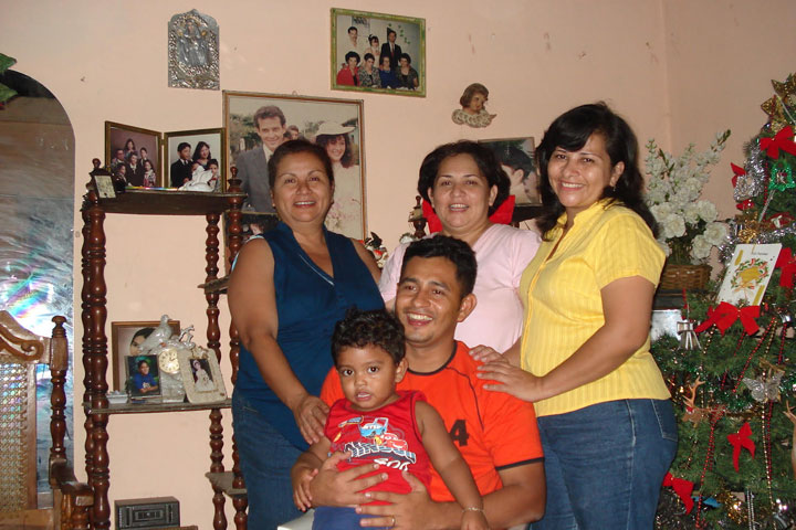Tres hermanas están de pie detrás de su hermano sentado, que sostiene un bebé. Todos tienen una gran sonrisa.