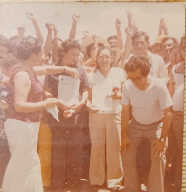 Una foto antigua, con un tono descolorido, de una celebración del Día del Maestro, que muestra a Efraín ligeramente inclinado mientras la gente al fondo levanta los puños y dos dedos en el aire. Junto a Efraín, dos personas muestran papeles de aspecto oficial a la cámara.