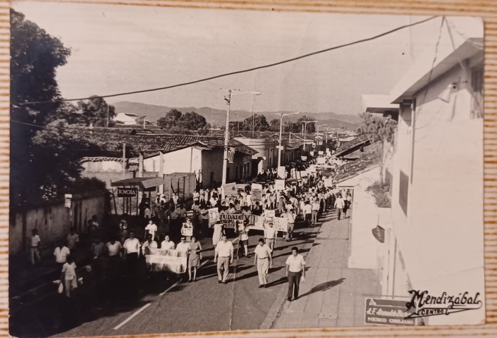 Una foto en blanco y negro de una manifestación en una calle de un pueblo, tomada desde un ángulo alto, mirando hacia abajo. La calle está llena de gente, a lo largo de toda la calle, presumiblemente más personas, marchando y sosteniendo pancartas. El sol de la tarde proyecta largas sombras.